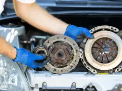 Mechanik w niebieskich rękawiczkach pokazuje zużyte części: sprzęgło i dwumasę. Dowiedz się, ile kosztuje wymiana sprzęgła i dwumasy.