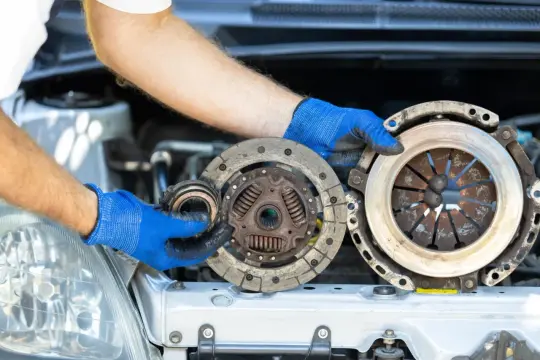 Mechanik w niebieskich rękawiczkach pokazuje zużyte części: sprzęgło i dwumasę. Dowiedz się, ile kosztuje wymiana sprzęgła i dwumasy.